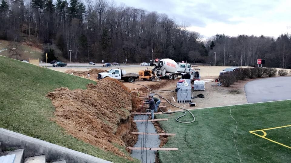 Construction site with workers pouring concrete; trucks and equipment near a stadium.