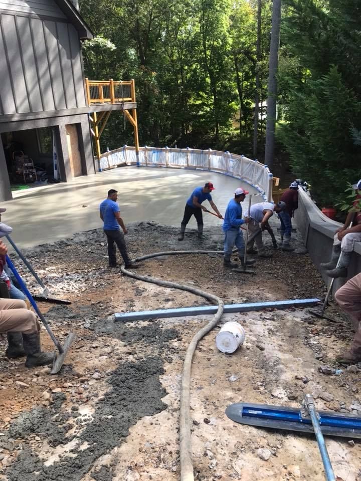 Construction workers pouring concrete on a curved surface outdoors. Several people using tools, with a building and trees in background.
