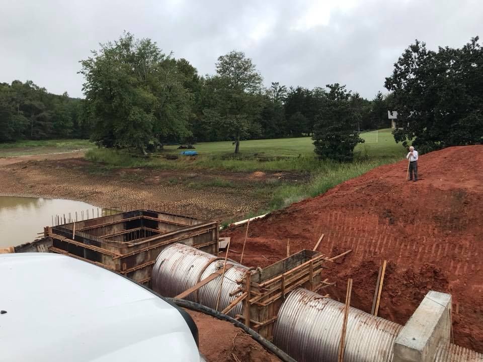 Construction site with corrugated pipes, red soil, and a person observing the work. Green field in background.