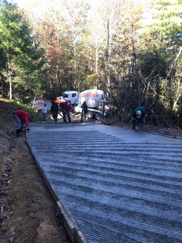 Construction workers pouring concrete on a roadway, with cement truck in background. Autumn trees line the sides.