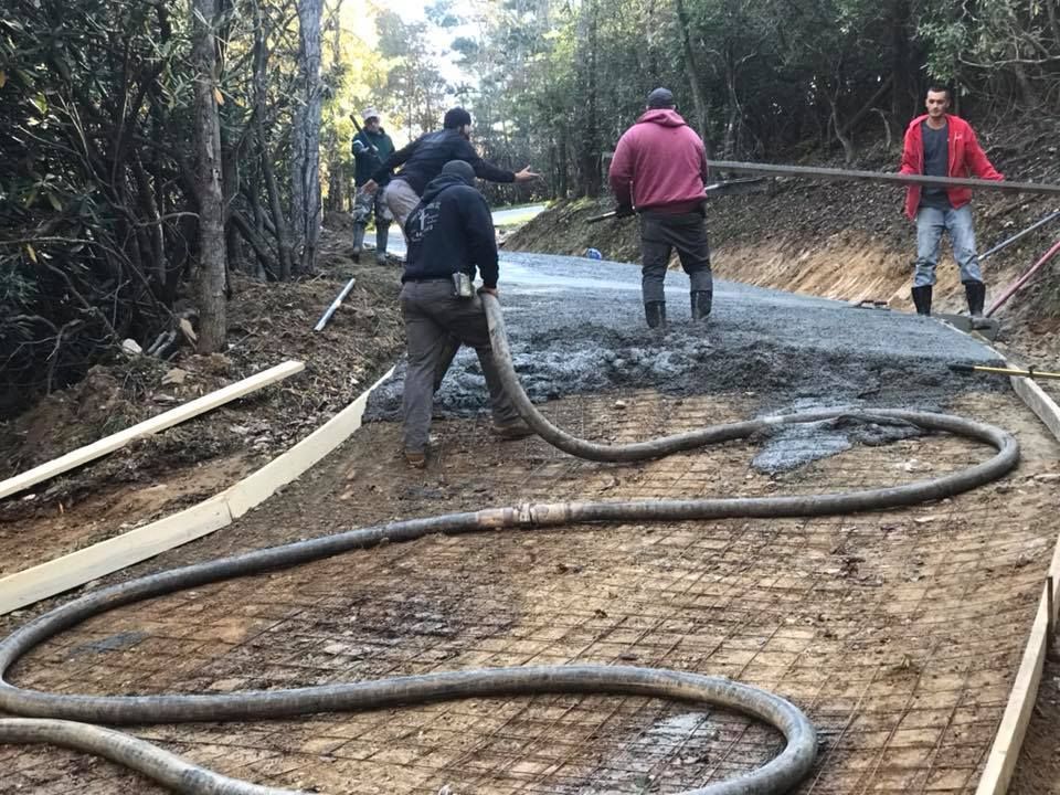 Workers pouring concrete on a road, using a hose. Forms and wire mesh visible. Forest background.