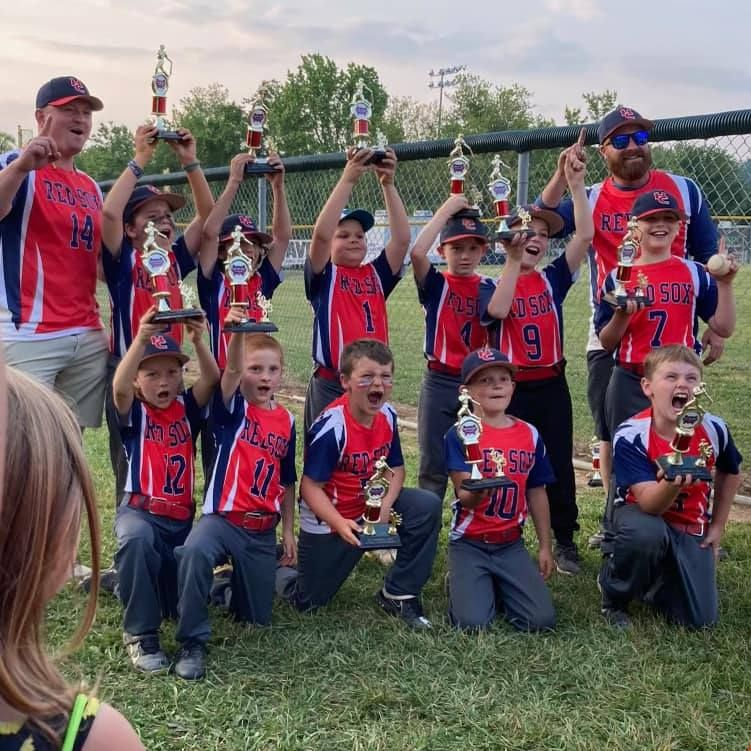 Youth baseball team celebrates victory, holding trophies. Team wearing red and blue uniforms in outdoor setting.