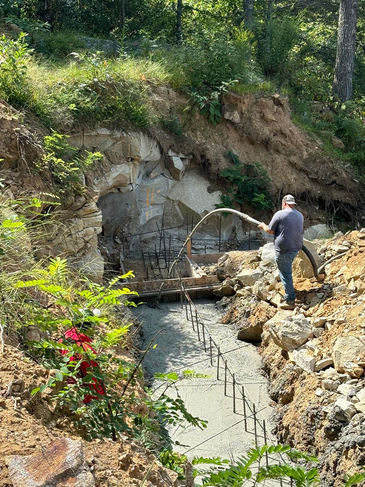 Man spraying concrete into a cave-like structure under construction, surrounded by dirt, trees, and greenery.