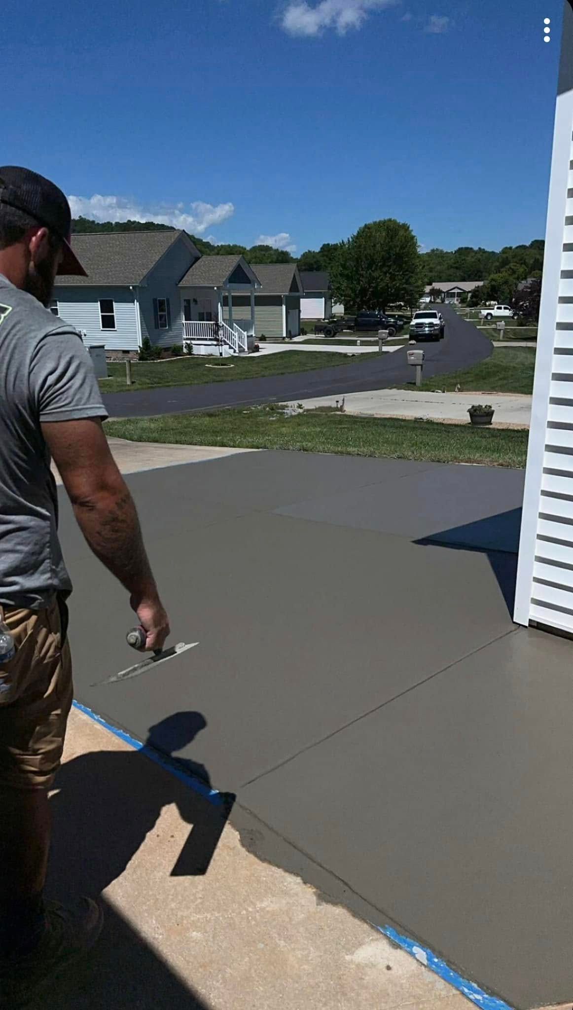 Man smoothing wet concrete on a patio with a trowel, sunny day, houses in the background.