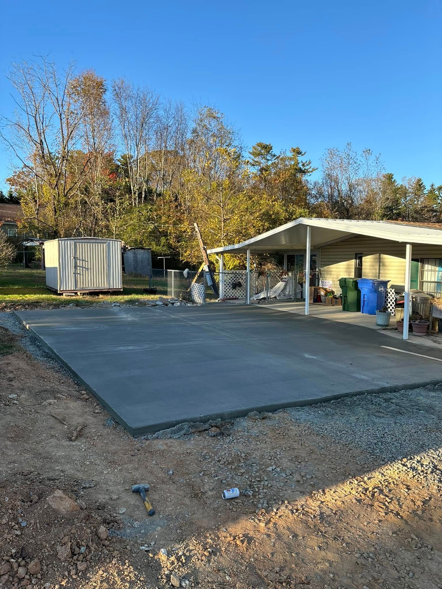 Concrete slab in front of a covered carport; gravel border; storage shed in the background.