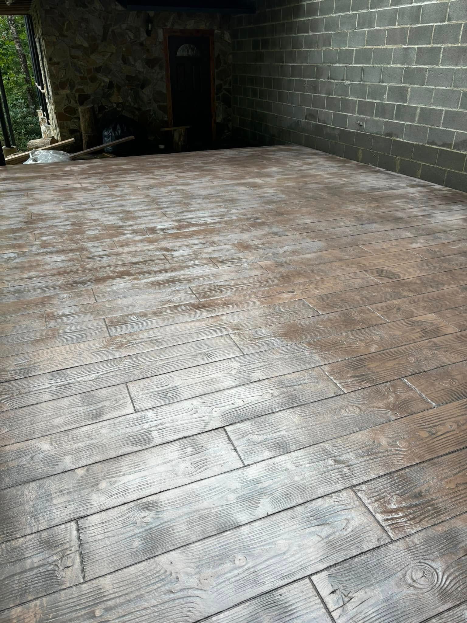 Stamped concrete patio with wood grain texture and gray-brown coloring. Adjacent to a brick wall.