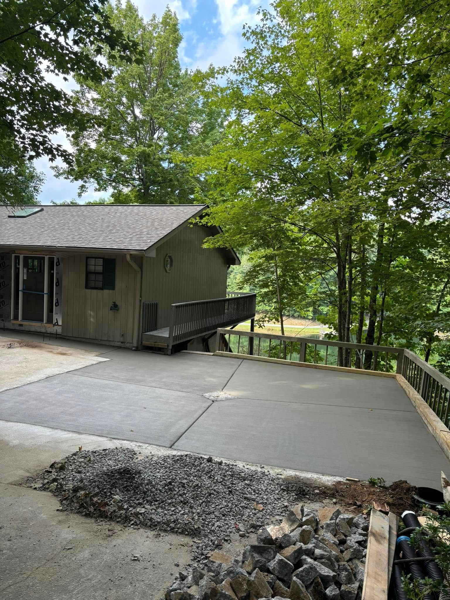 A house with a concrete patio. Trees and a deck with a railing overlook a body of water.