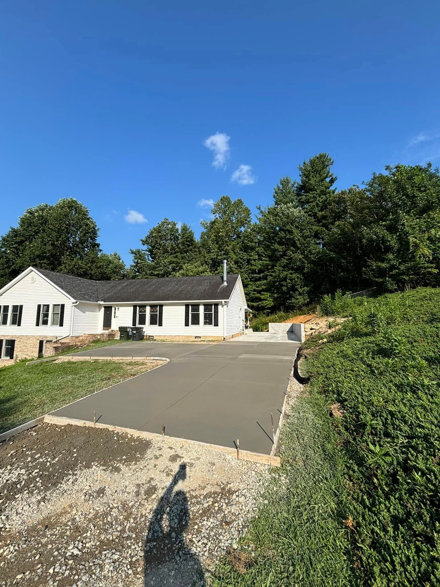 Freshly poured concrete driveway in front of a white house with black shutters, trees in the background, blue sky.