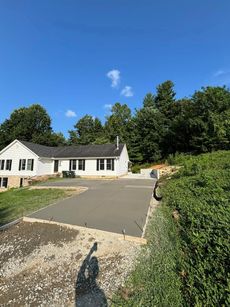 Freshly poured concrete driveway in front of a white house with black shutters, trees in the background, blue sky.
