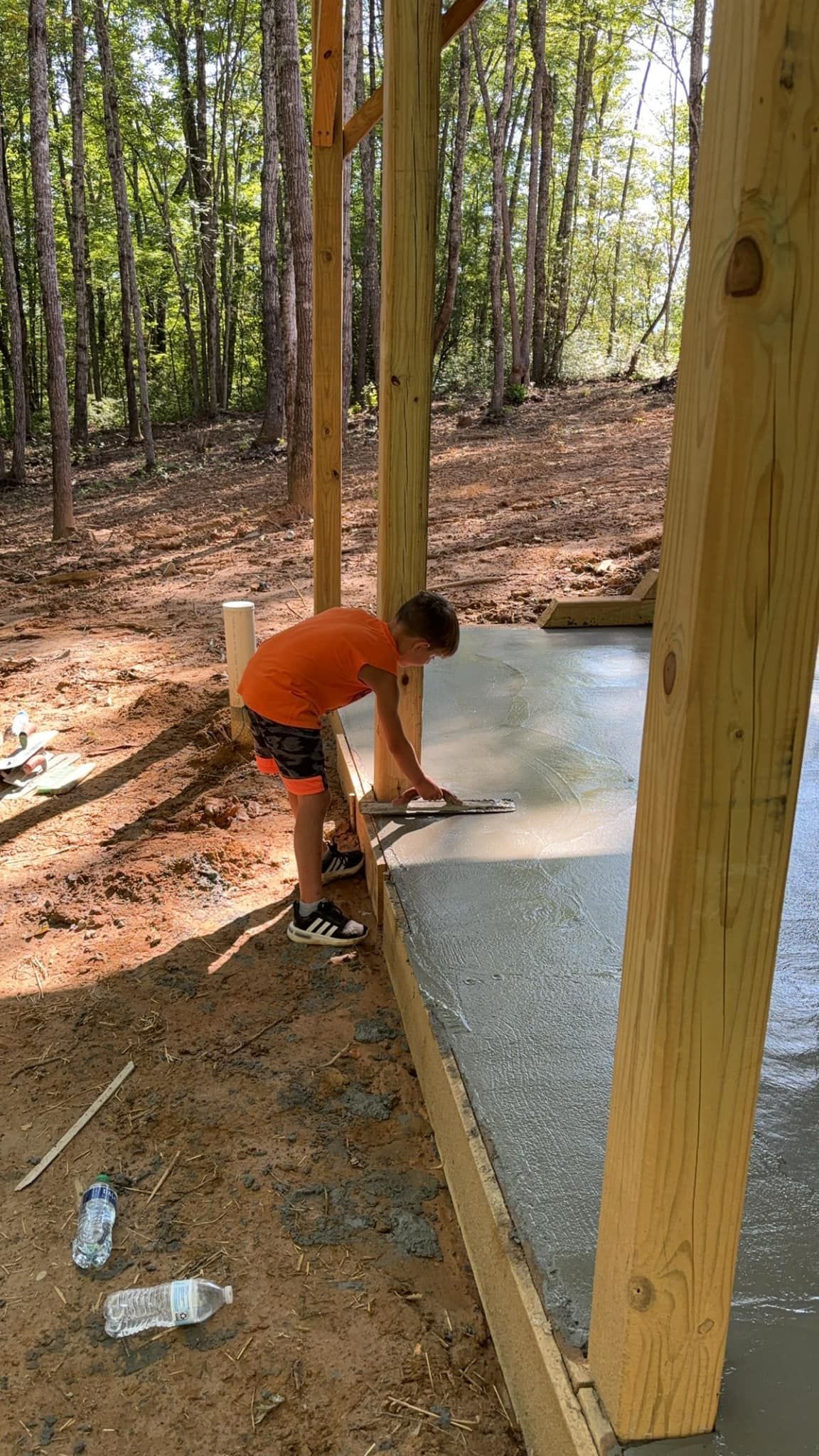 Person smoothing concrete, surrounded by wooden beams and trees.
