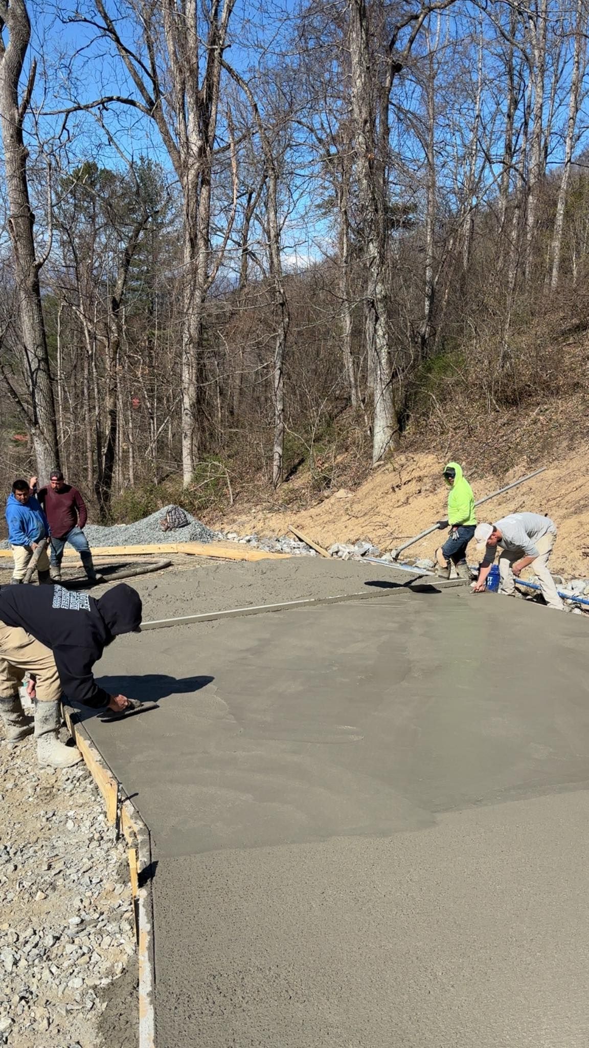 Construction workers pouring and smoothing concrete on a sunny day in a wooded area.