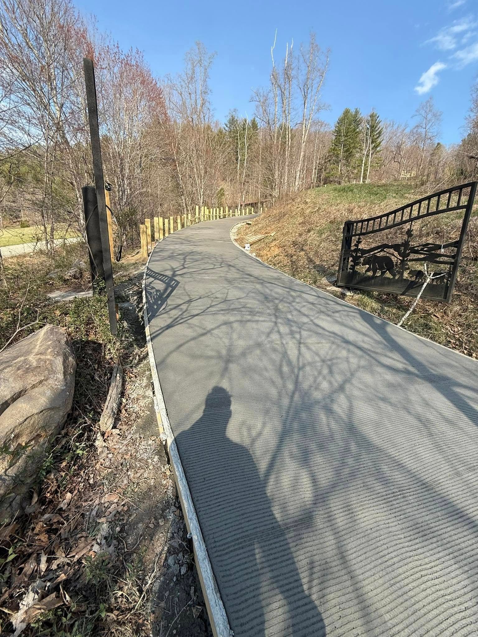 A paved pathway curves through a park, with trees on either side. Sunlight casts shadows on the path.