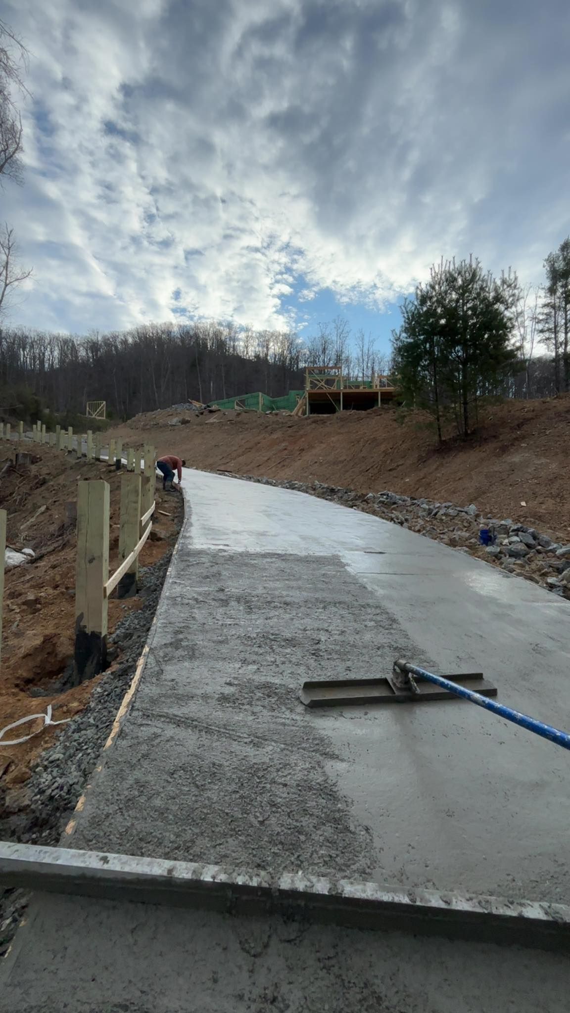 Concrete walkway construction in progress; workers smoothing the wet surface with a long-handled tool under a cloudy sky.