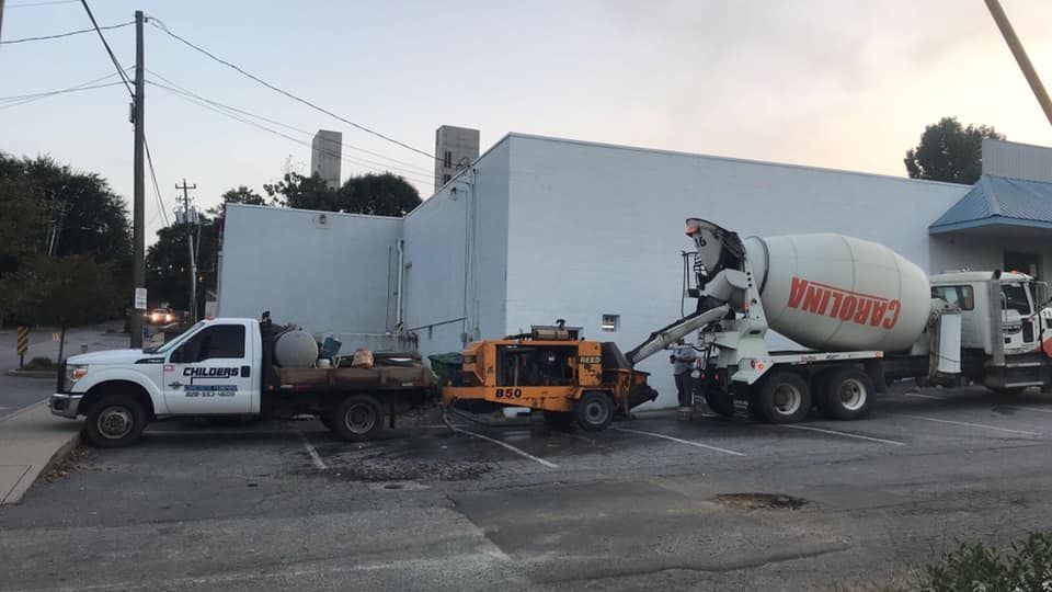 Concrete truck pouring cement in a parking lot next to a white building; other construction vehicles present.