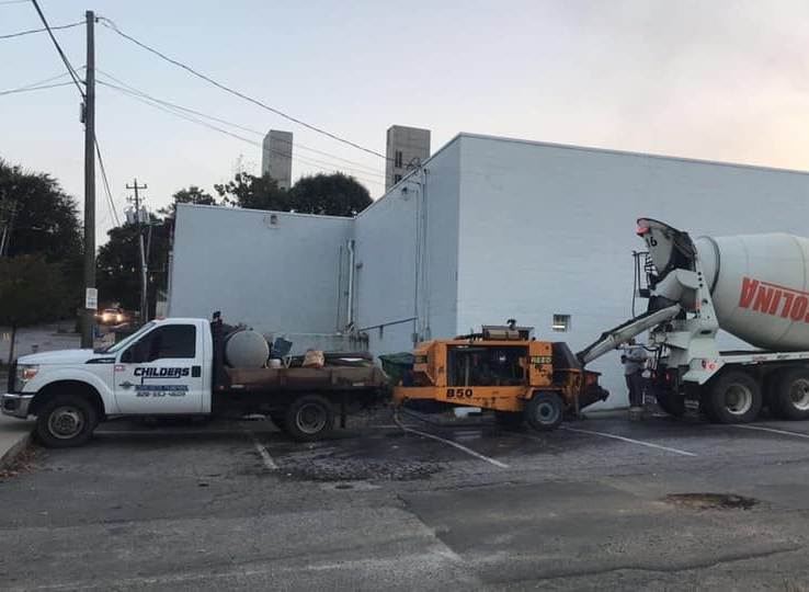 Concrete being poured from a truck into a pump on a parking lot next to a white building.