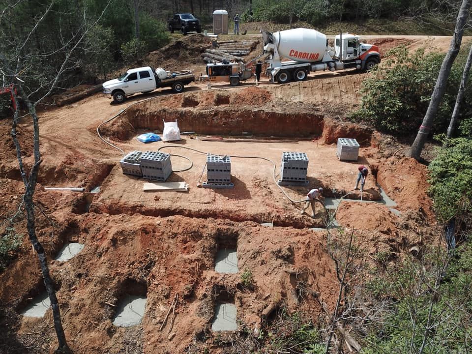 Construction site: truck pouring concrete into foundation, workers, surrounded by dirt and trees.