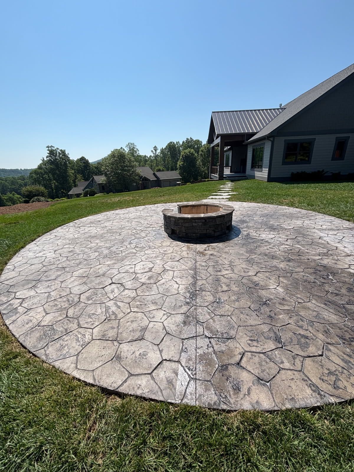 Circular stone patio with fire pit, grassy yard, and house under a blue sky.