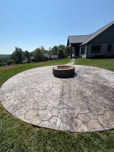 Circular stone patio with fire pit, grassy yard, and house under a blue sky.