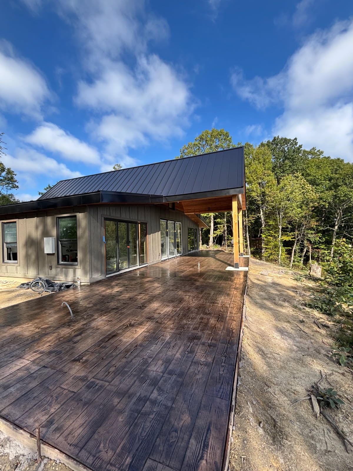 Modern cabin with dark metal roof, large wooden deck, and surrounding trees under a blue sky.