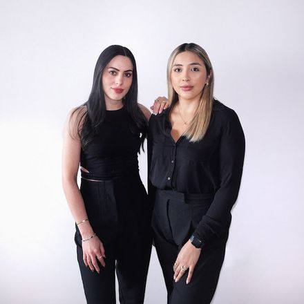 Two women in black outfits standing against a light studio backdrop