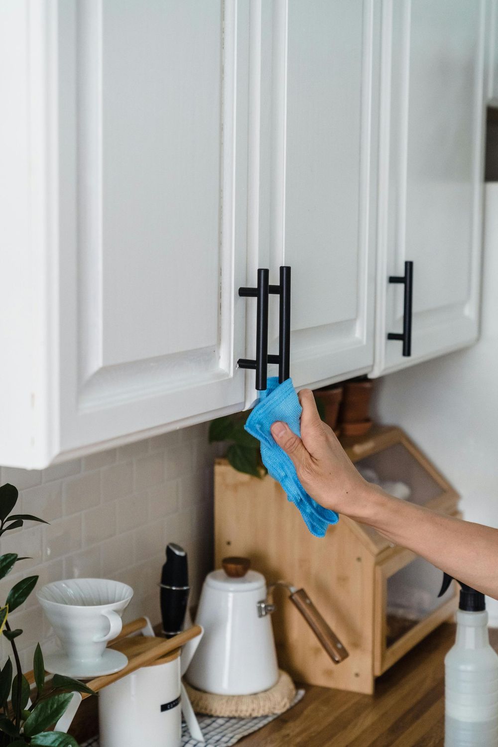 A hand holding a blue cloth cleans a white kitchen cabinet door with a black handle.