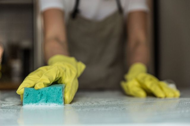 A person wearing yellow rubber gloves wipes a white surface with a teal sponge.
