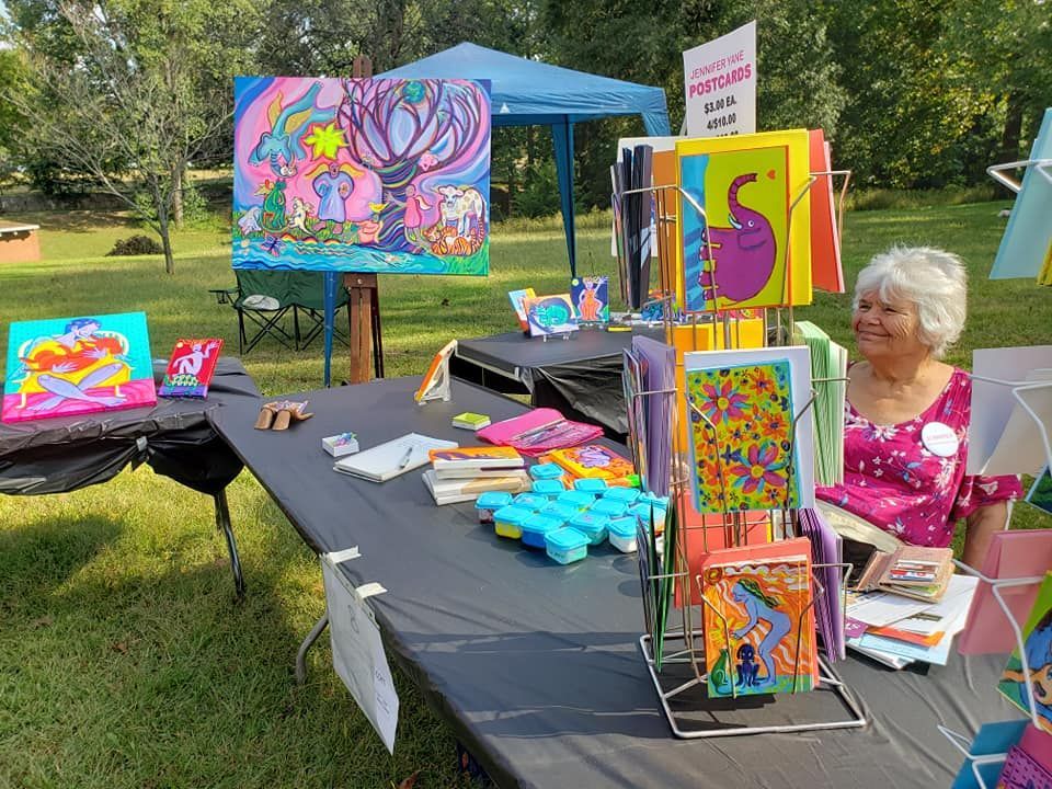 Art fair booth with woman, colorful paintings, postcards, and small items on display in a park setting.