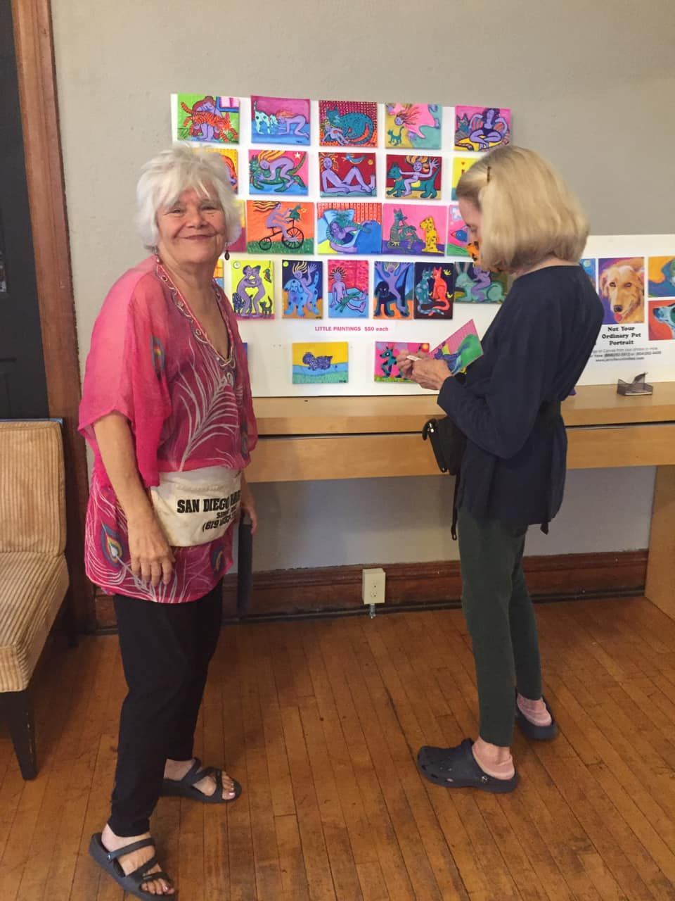Two women at an art display; one smiles at the viewer, the other examines the colorful paintings.