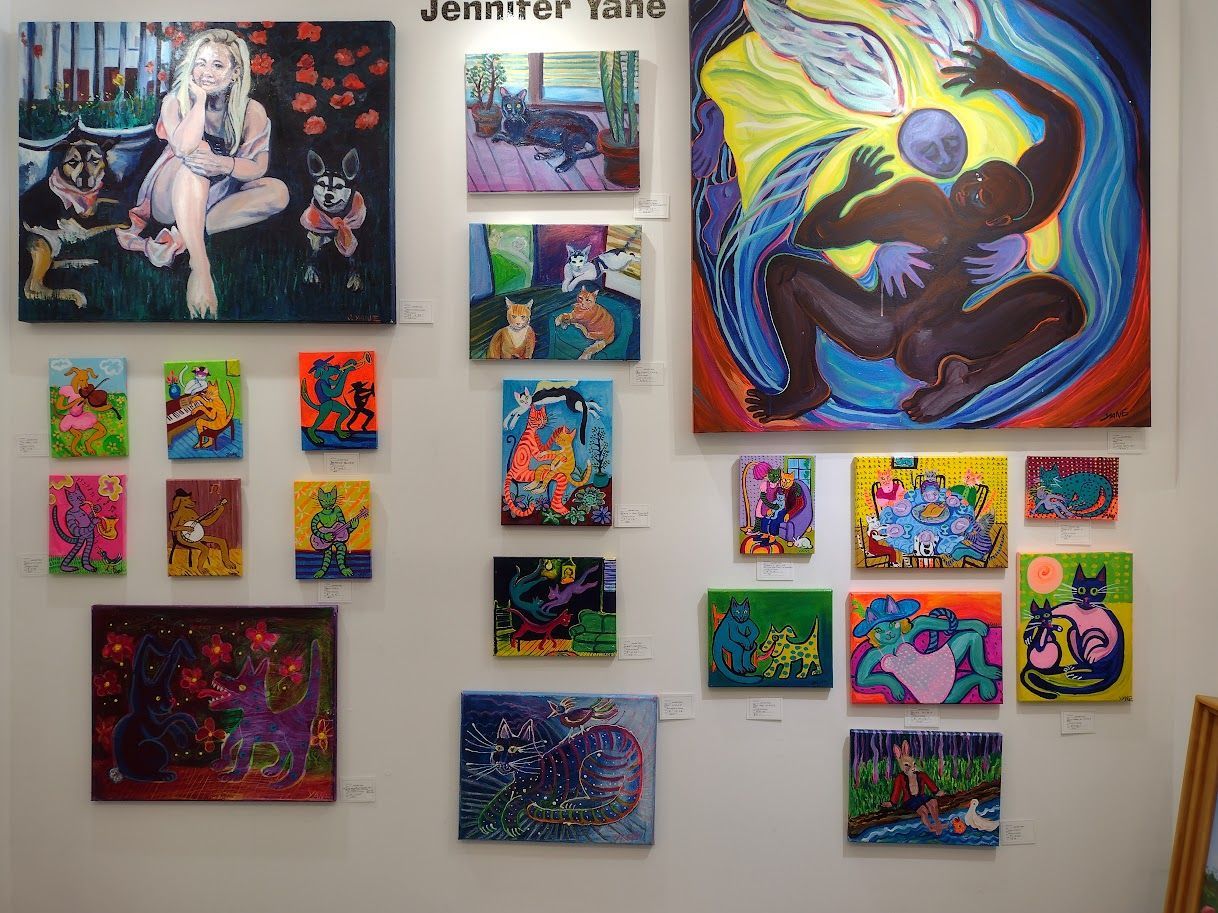 Artwork display: Colorful paintings of figures, animals, and abstract scenes hang on a white wall in a gallery setting.