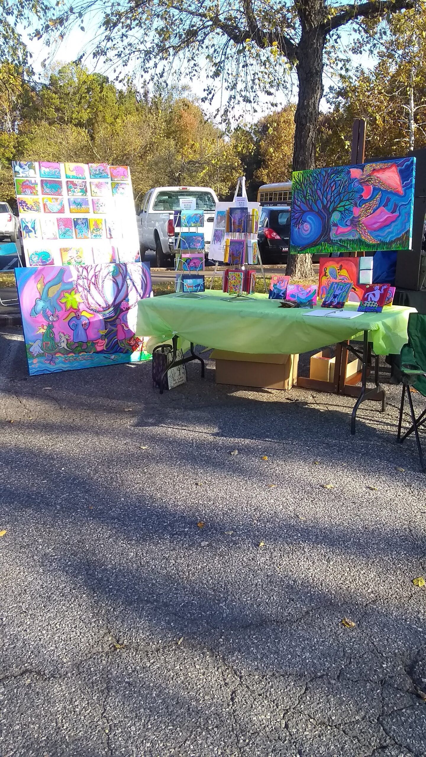 An outdoor art display with colorful paintings on a table and leaning against a structure on a gravel lot.