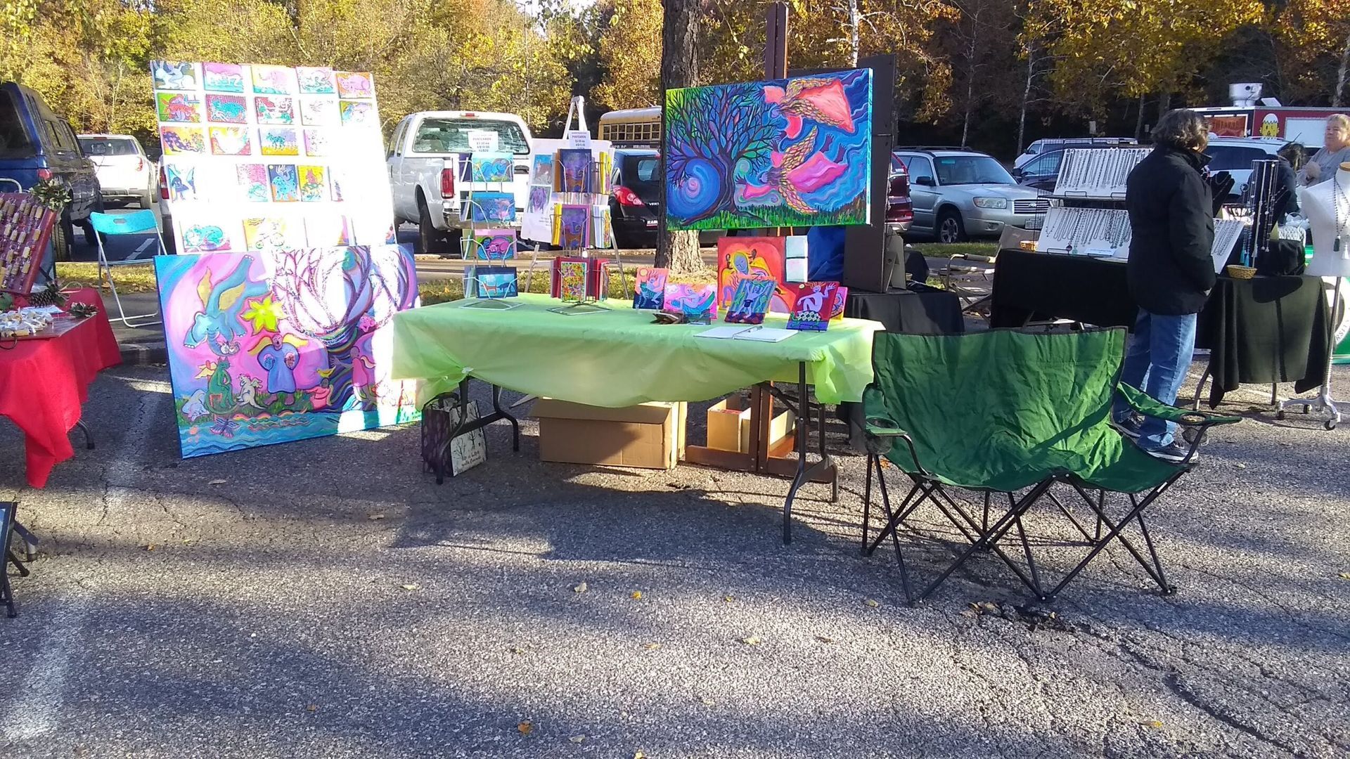 Art booth at an outdoor market with paintings, various colors, a green table, and a green folding chair.