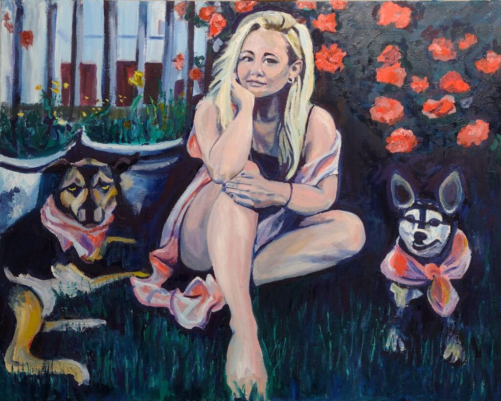 Woman and two dogs sit in a garden. The woman is blonde, the dogs have bandanas on.