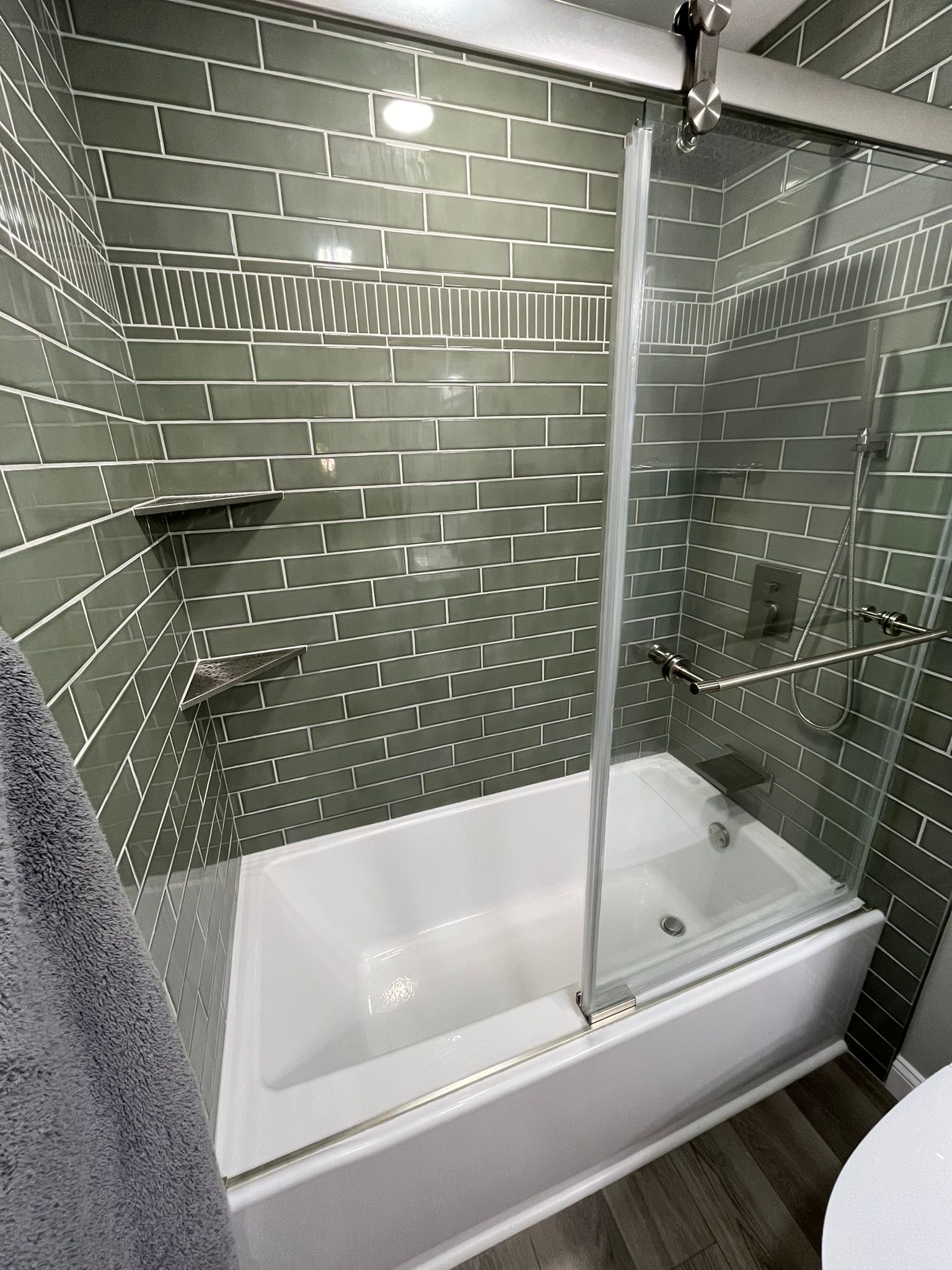 A modern bathroom with green tiled walls, a white tub, and a glass shower door.