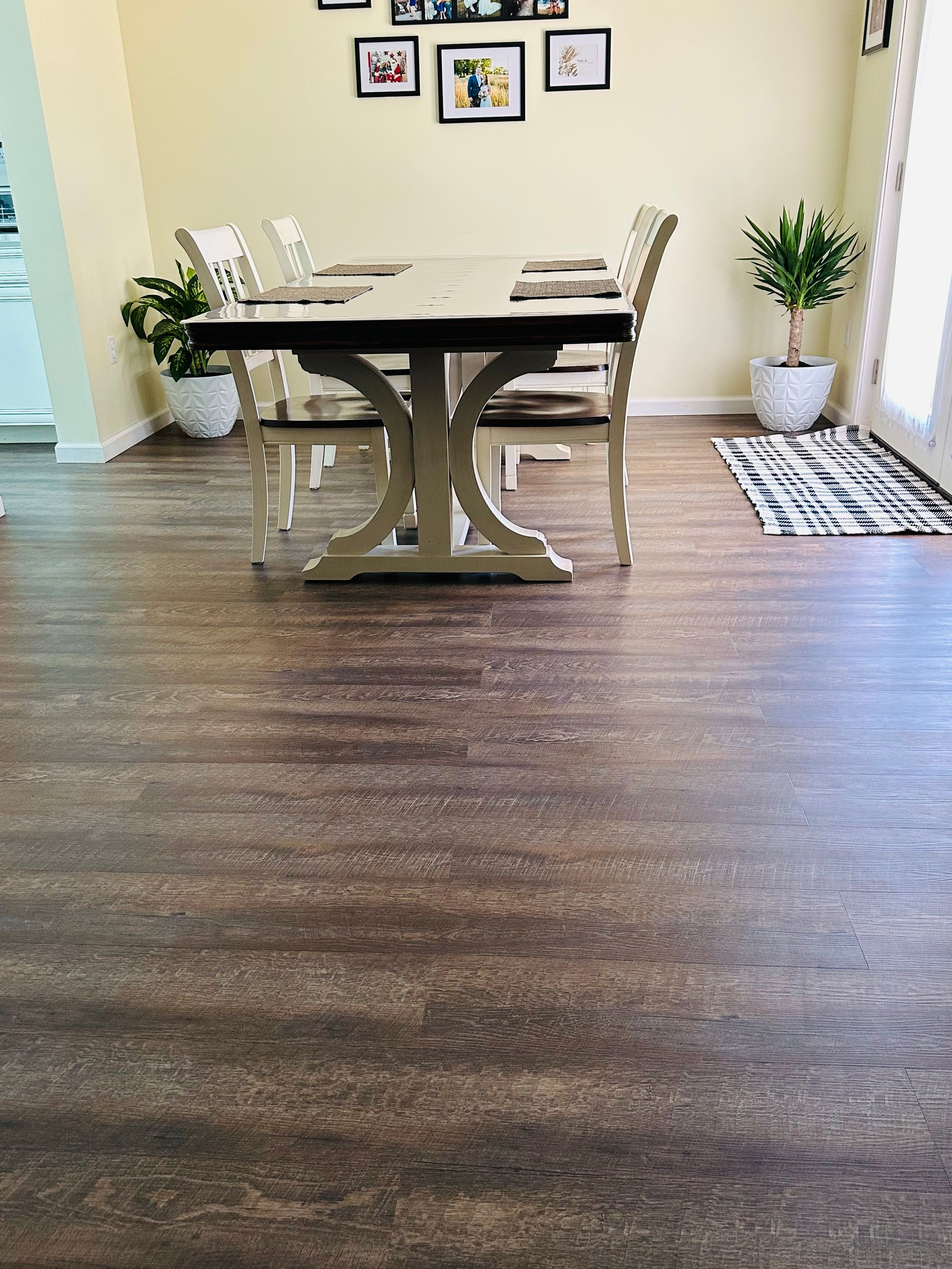 Dining room with a light wooden table and chairs, dark wood-look flooring, and a few decorative plants.