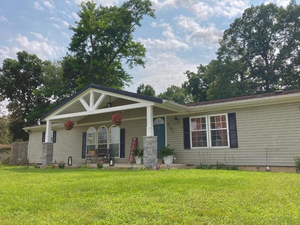 Tan house with a covered porch, green lawn, and trees against a cloudy sky.