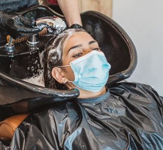Young woman have hair cutting at hair stylist during pandemic isolation, they both wear protective equipment. Hair stylist washing her hair