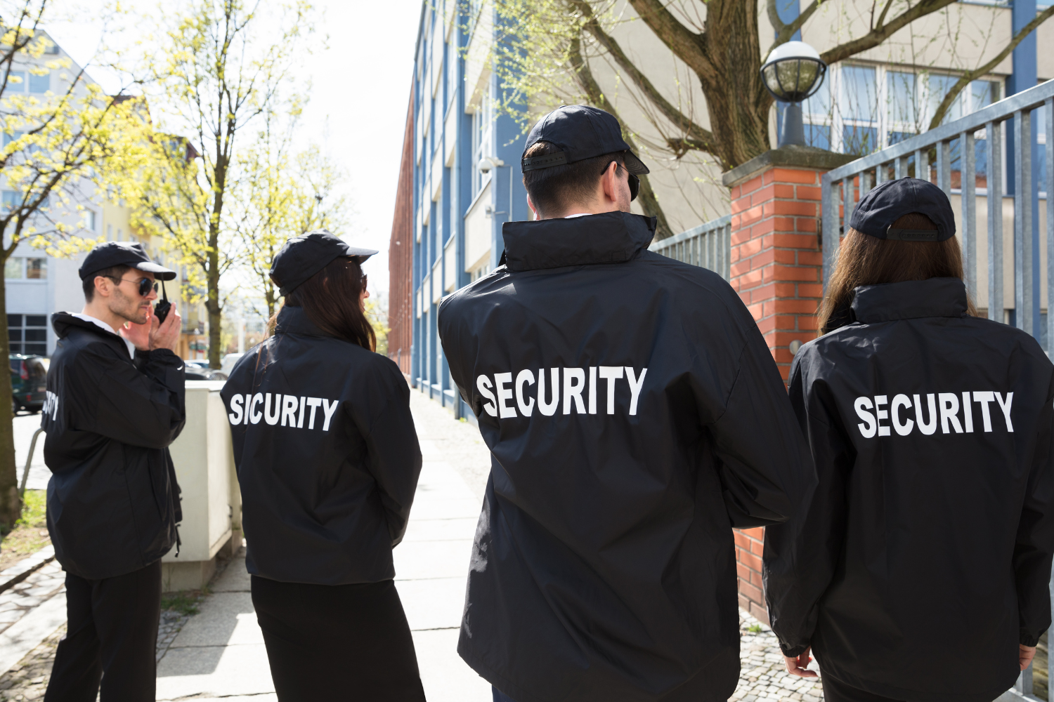 Four security guards in black jackets standing outside a building. One uses a radio. 