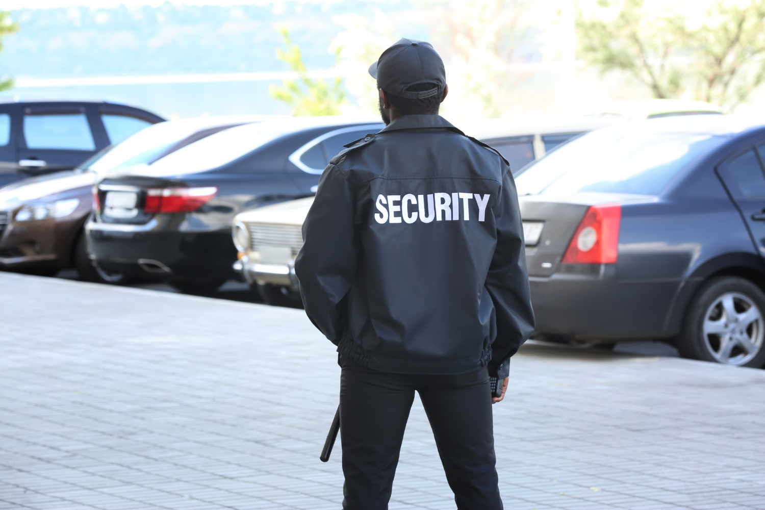 Security guard in black uniform stands in a parking lot, facing away from the camera.