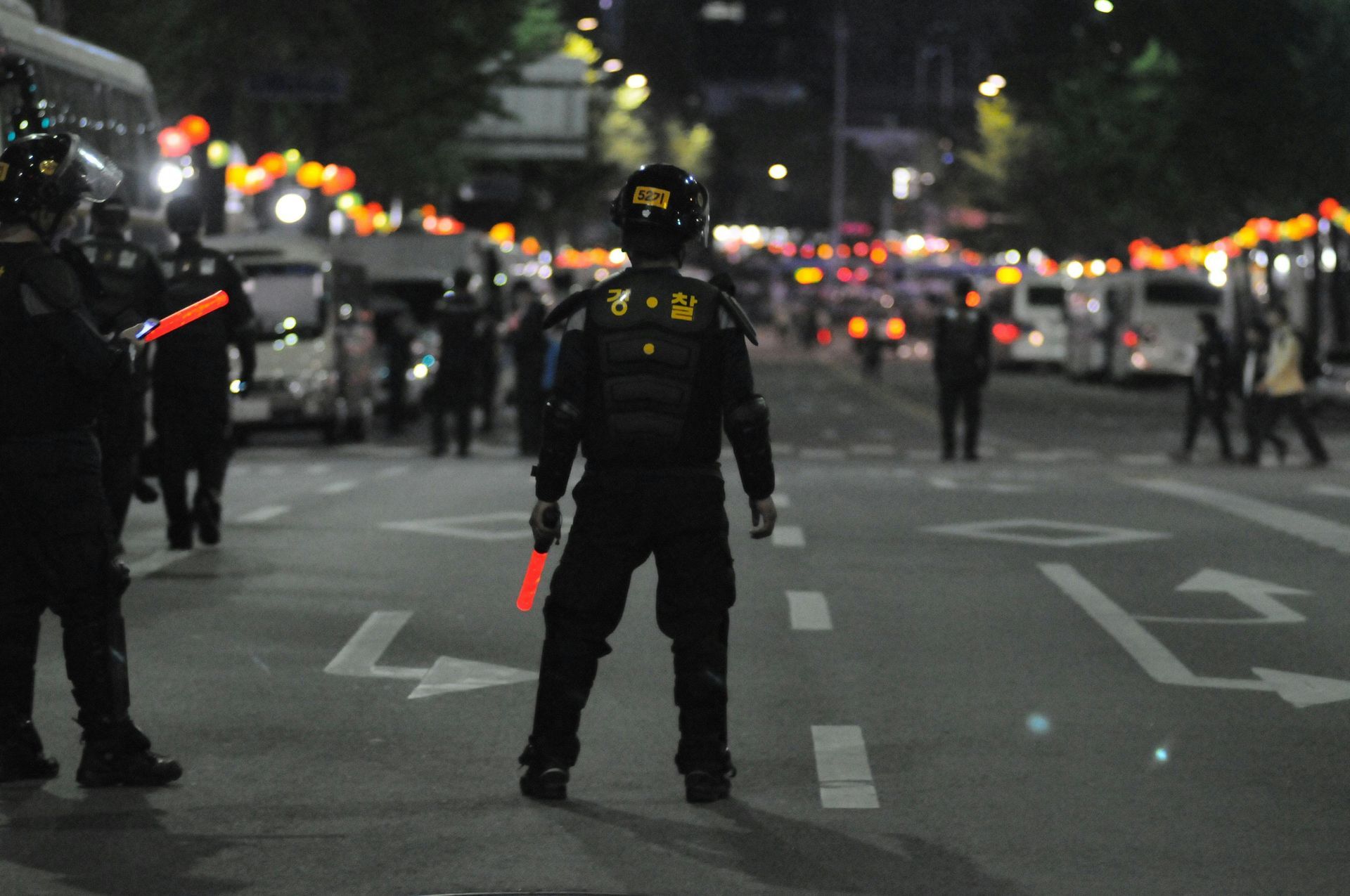 Police officers in riot gear stand on a city street at night, with vehicles and people in the background.