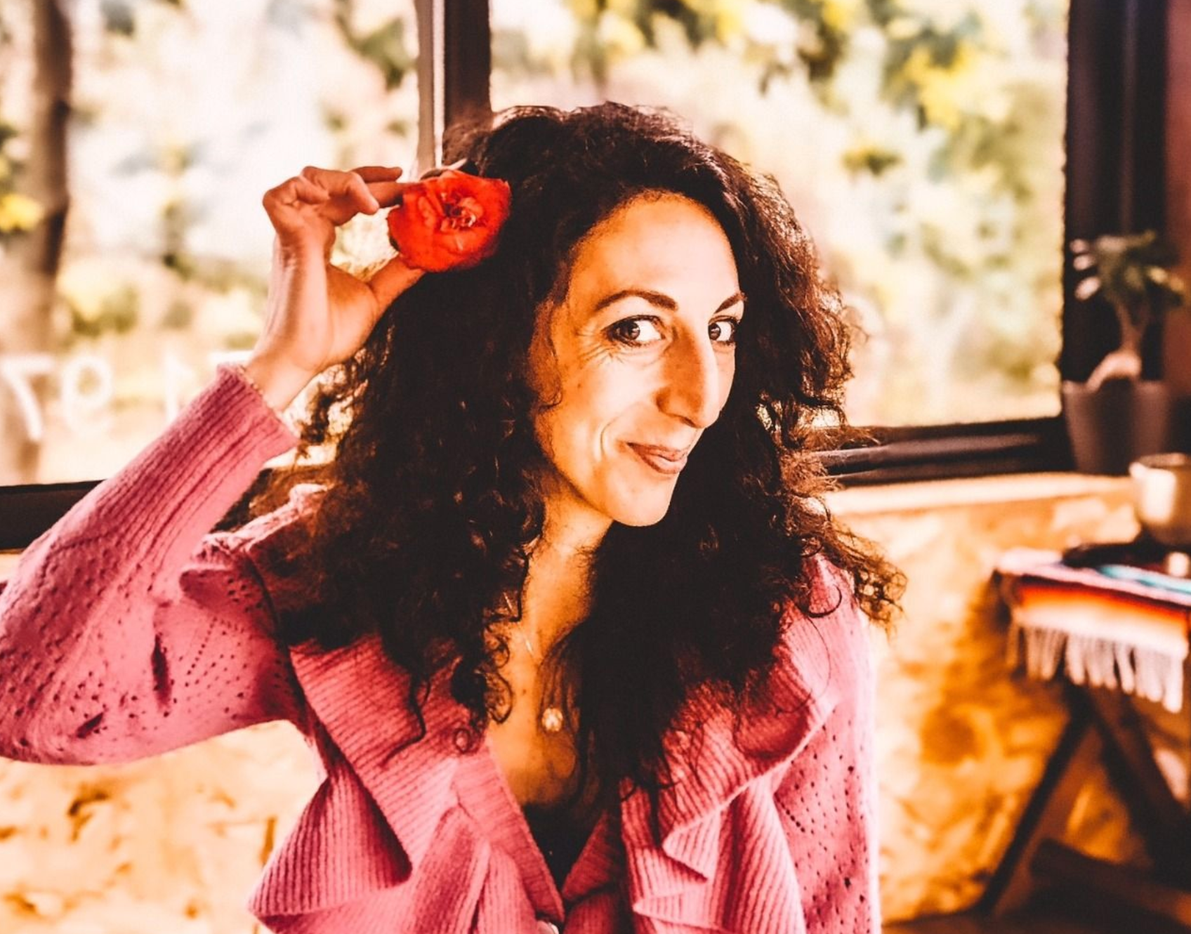 Une femme vêtue d'un pull rose tient une fleur rouge dans ses cheveux.