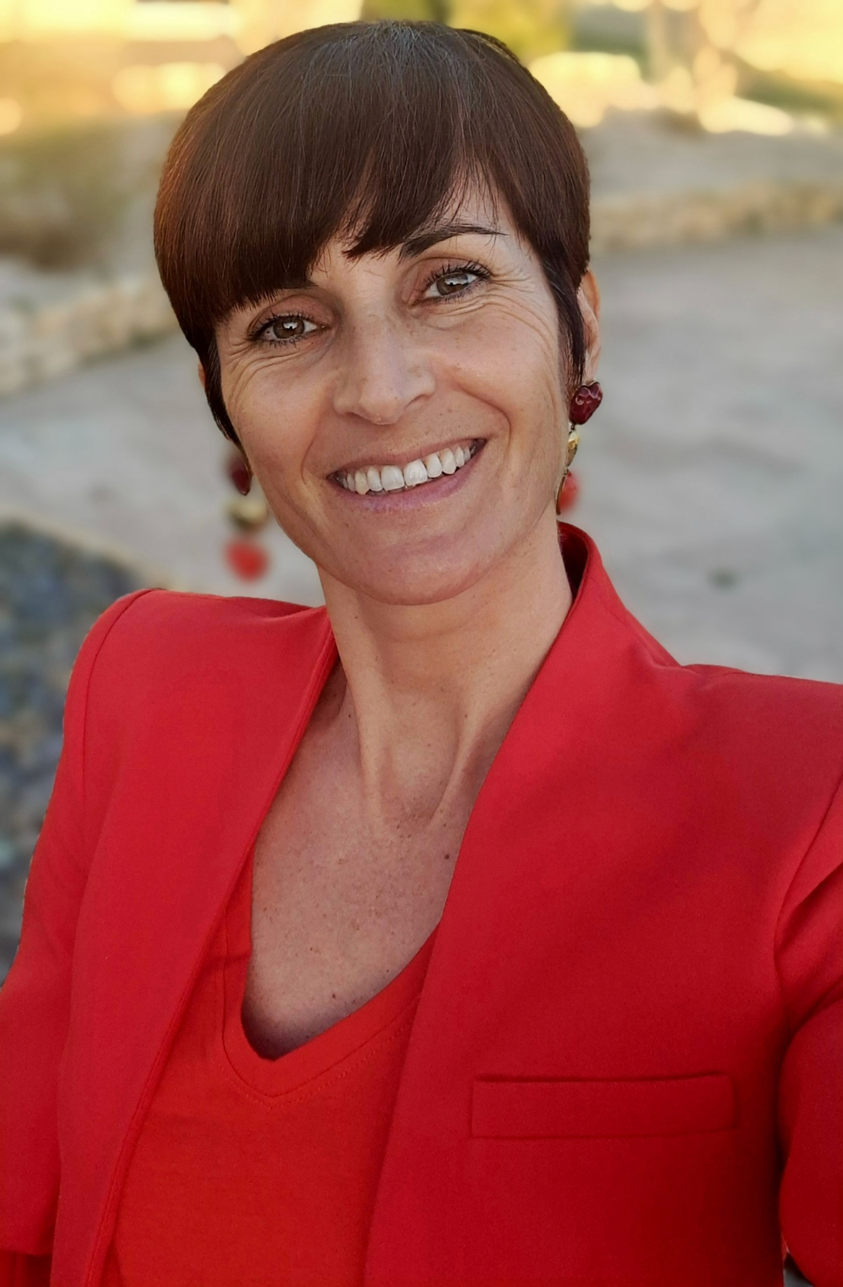 Une femme portant une veste rouge et des boucles d'oreilles sourit à la caméra.