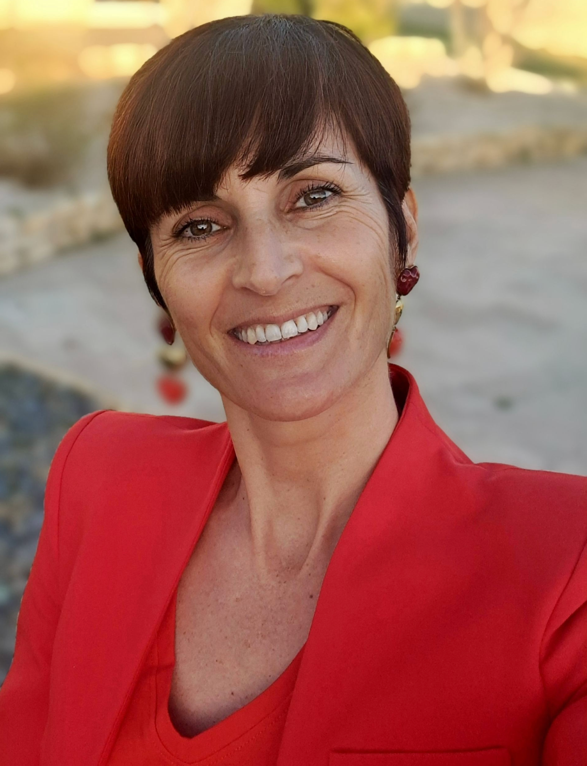 Une femme portant une veste rouge et des boucles d'oreilles sourit à la caméra.