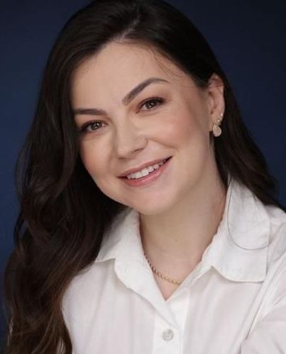 Uma mulher com longos cabelos castanhos e uma jaqueta branca está sorrindo para a câmera.