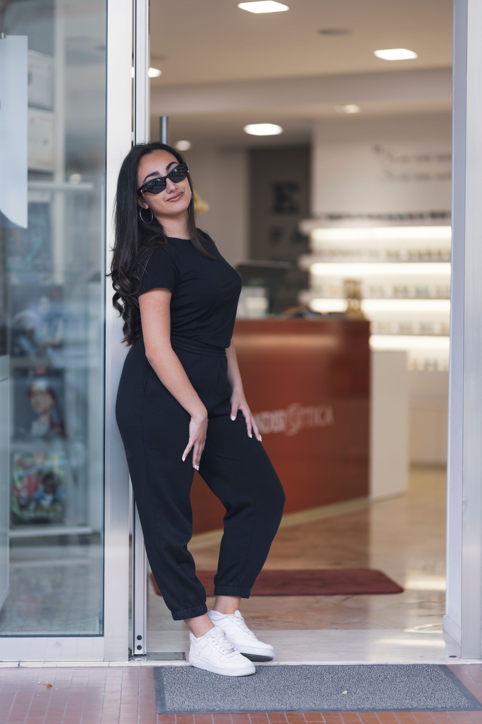 Una donna con una tuta nera e delle scarpe da ginnastica bianche è appoggiata a una porta a vetri.