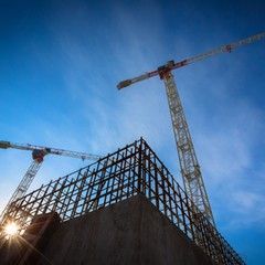 Construction site with two cranes, a building frame, and a bright sun against a blue sky.