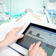 Hands operating a medical ventilator, displaying a patient's breathing data, with a patient in the background.