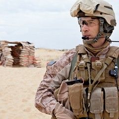 Soldier in combat gear, helmet and vest, surveys desert landscape near a makeshift shelter.
