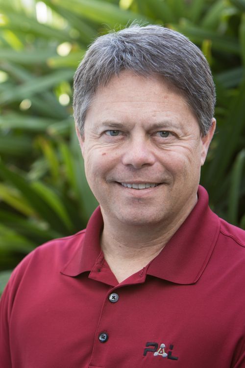 Woman with glasses smiling, wearing a black and white patterned shirt, and a pendant necklace, in front of greenery.