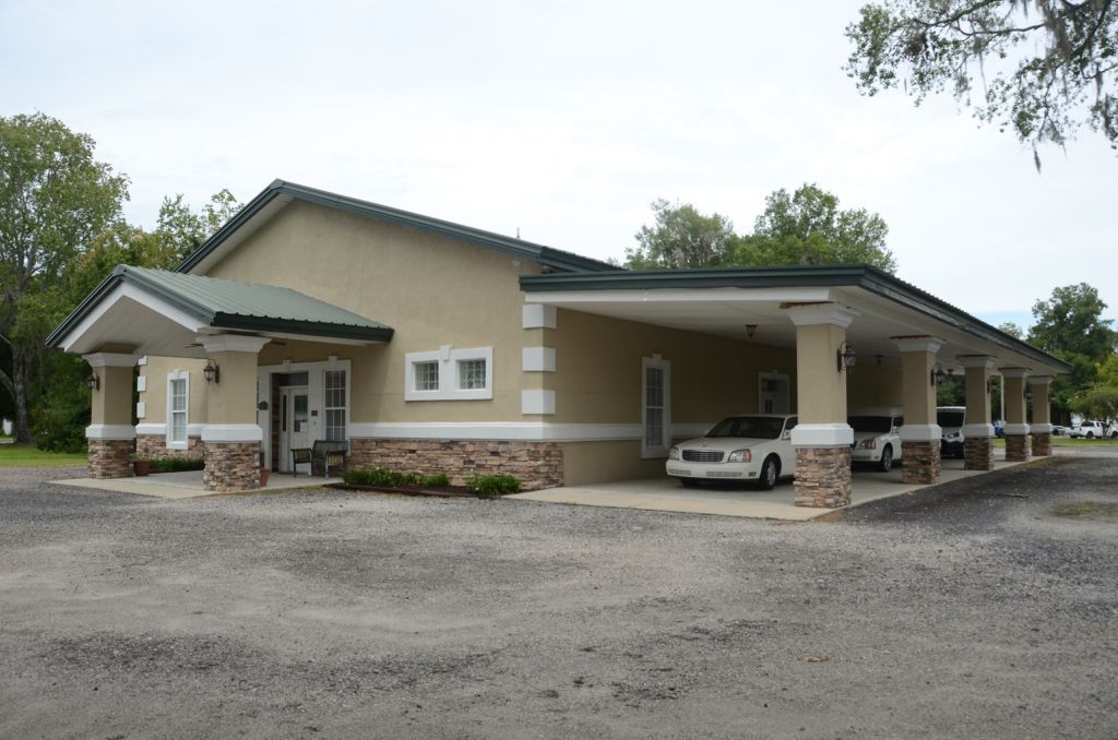 A light green building with a porch and a large asphalt parking lot. A car and a hearse are parked there.