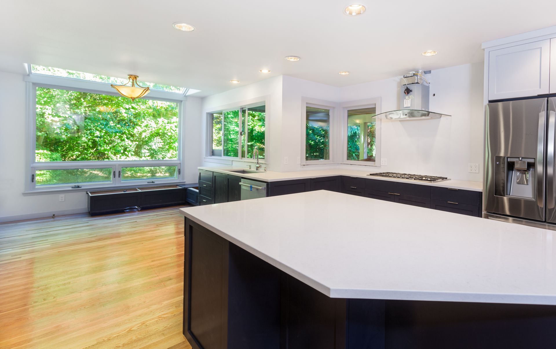 Modern kitchen with white countertops, dark cabinets, and large windows overlooking trees.